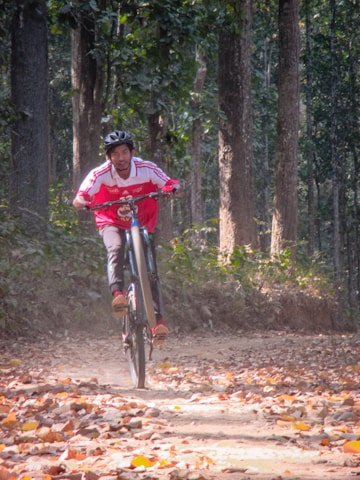 A person wearing a red and white sports jersey and a helmet rides a mountain bike through a forested trail. The path is covered with dry leaves and surrounded by tall trees with green foliage. The rider is doing a wheelie, balancing the bike on its back wheel.