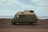 A green camper van is parked on a rocky beach with the ocean in the background. The landscape is desolate with scattered vegetation, and the sky is partly cloudy. The van has a roof cargo holder and appears to be ready for travel or camping.