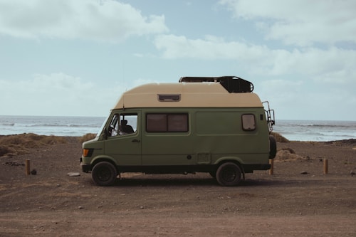 A green camper van is parked on a rocky beach with the ocean in the background. The landscape is desolate with scattered vegetation, and the sky is partly cloudy. The van has a roof cargo holder and appears to be ready for travel or camping.