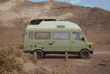 A light green camper van is parked on a dirt path in front of a rugged, rocky mountain landscape. The van has a boxy shape with windows and a roof vent, and it appears to be an older model with a retro design. The surrounding environment is arid, with sparse bushes and gravel.