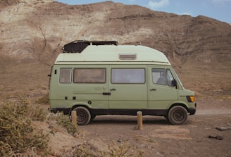 A light green camper van is parked on a dirt path in front of a rugged, rocky mountain landscape. The van has a boxy shape with windows and a roof vent, and it appears to be an older model with a retro design. The surrounding environment is arid, with sparse bushes and gravel.
