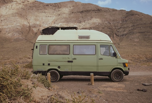 A light green camper van is parked on a dirt path in front of a rugged, rocky mountain landscape. The van has a boxy shape with windows and a roof vent, and it appears to be an older model with a retro design. The surrounding environment is arid, with sparse bushes and gravel.