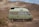 A light green camper van is parked on a dirt path in front of a rugged, rocky mountain landscape. The van has a boxy shape with windows and a roof vent, and it appears to be an older model with a retro design. The surrounding environment is arid, with sparse bushes and gravel.