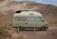A light green camper van is parked on a dirt path in front of a rugged, rocky mountain landscape. The van has a boxy shape with windows and a roof vent, and it appears to be an older model with a retro design. The surrounding environment is arid, with sparse bushes and gravel.