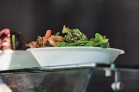 A white disposable plate contains a fresh salad with green leafy vegetables and crispy fried pieces, possibly croutons or fried onions. The plate is placed on a glass surface, and there is soft lighting creating a reflective effect. The background is blurred, emphasizing the foreground dish.