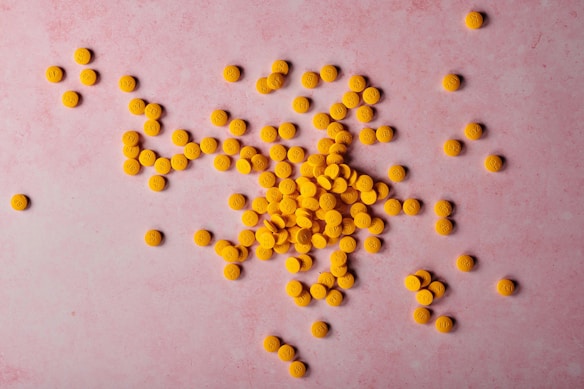 A scattered group of small, orange, round tablets is spread across a pink surface. Each tablet has imprints on one side, featuring numbers and letters.