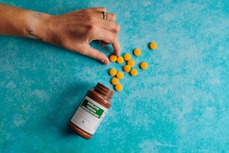 A hand is reaching for a pile of yellow tablets scattered next to an open brown bottle on a blue textured surface. The bottle likely contains dietary supplements or medication, as indicated by the label.