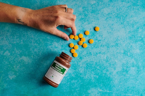 A hand is reaching for a pile of yellow tablets scattered next to an open brown bottle on a blue textured surface. The bottle likely contains dietary supplements or medication, as indicated by the label.