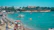 Tourists sipping drinks and relaxing on lounge chairs at a sunny beach club by the turquoise sea.