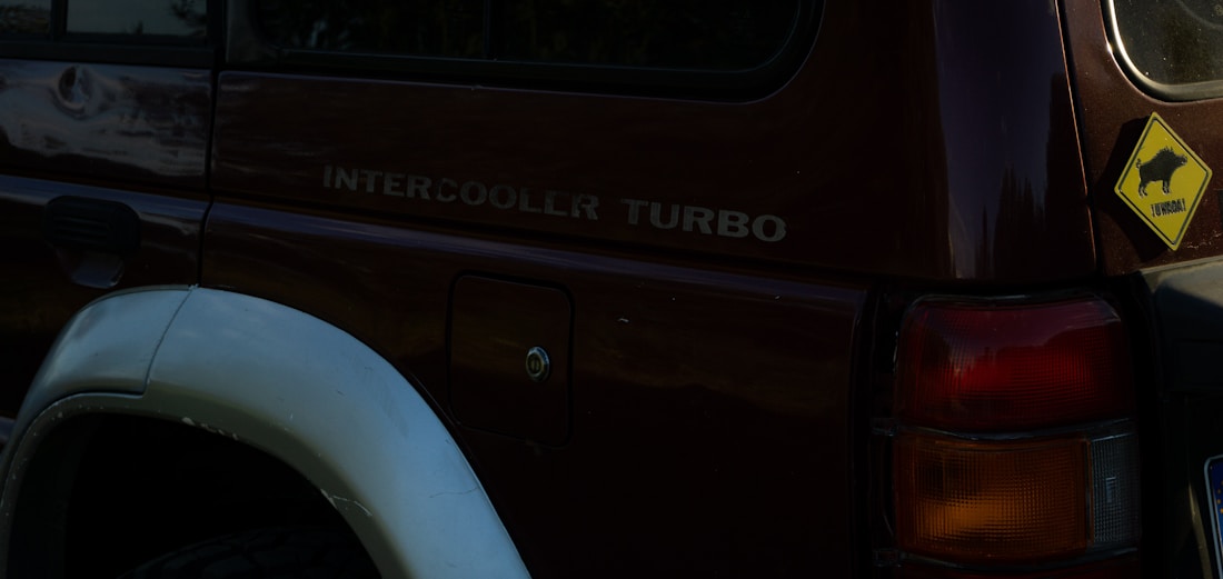 A dark-colored vehicle with visible text 'INTERCOOLER TURBO' on its side and a yellow caution sign featuring an outline of a buffalo, with the word 'hombare!' beneath it. The vehicle is parked, with part of its wheel arch and indicator light visible.