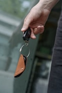 A hand holding a car key fob attached to a brown leather key holder in front of a blurred background.