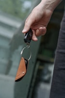 A hand holding a car key fob attached to a brown leather key holder in front of a blurred background.