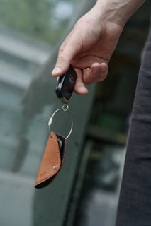 A hand holding a car key fob attached to a brown leather key holder in front of a blurred background.
