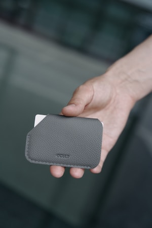 A hand holds a textured gray cardholder embossed with 'CULT' on its surface. A white card is partially visible, protruding from the top of the cardholder. The background is blurred, featuring muted shades of gray and green.