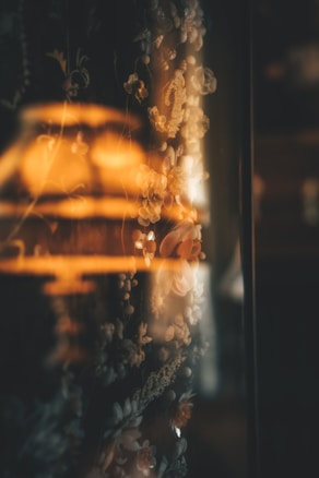 A warm, dimly lit scene with a floral patterned fabric in the foreground. The fabric has intricate details of flowers and vines with soft focus, creating a cozy and intimate atmosphere. The warm glow of a lamp light reflects off the fabric, enhancing the floral patterns.