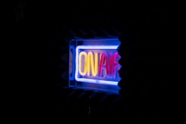 A glowing neon sign displaying the words 'ON AIR' in vibrant colors against a dark background. The surrounding area appears to be in a studio setting with soundproofing foam visible.