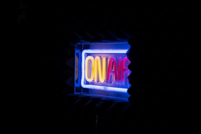 A glowing neon sign displaying the words 'ON AIR' in vibrant colors against a dark background. The surrounding area appears to be in a studio setting with soundproofing foam visible.