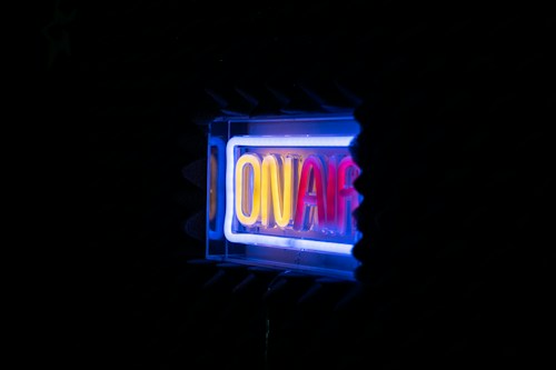 A glowing neon sign displaying the words 'ON AIR' in vibrant colors against a dark background. The surrounding area appears to be in a studio setting with soundproofing foam visible.