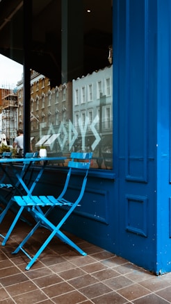 A small outdoor seating area with two blue metal chairs and a matching table is seen against a vibrant blue storefront. The window reflects nearby buildings and street activity, while large, stylized text is visible on the glass.
