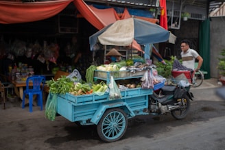 A small motorized cart carrying an assortment of fresh vegetables and produce is parked on a street. The cart is blue and has a canopy. Various vegetables like leafy greens, cucumbers, and cabbages are visible. A man is seen nearby, with a shop in the background displaying hanging bags and goods. There is a blue plastic chair next to the cart, and the scene conveys a typical street market or vendor setting.