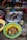 Various bowls and basins are filled with live fish and eels, displayed on the ground in an outdoor market setting. A person wearing green boots is seated nearby, and there is a scale and other market paraphernalia in the background.