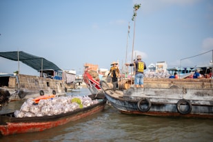 Two boats on a river are engaged in trade, with one loaded with bags of goods and people standing on the deck. The bustling scene suggests a lively market atmosphere. Nearby, other boats and buildings create a backdrop under a clear blue sky.