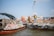 Two boats on a river are engaged in trade, with one loaded with bags of goods and people standing on the deck. The bustling scene suggests a lively market atmosphere. Nearby, other boats and buildings create a backdrop under a clear blue sky.