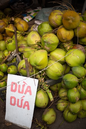 A large pile of green coconuts with thin husks is stacked at a market stall. In front of the pile, there is a sign with red writing on it, suggesting they might be for sale.