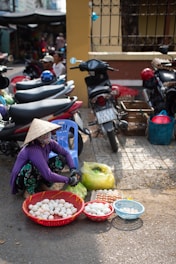 A street vendor squats next to baskets of eggs laid out for sale on a city sidewalk. The vendor wears a traditional conical hat, a face covering, and colorful clothing while seated on a blue plastic chair. Nearby are parked motorcycles and there is a building with a fence in the background.