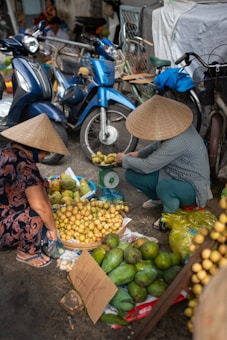 Two people wearing traditional conical hats are seen arranging and weighing yellow fruits at a street market. The scene is bustling with motorbikes and bicycles parked in the background. The ground is cluttered with various fruits, plastic bags, and cardboard.