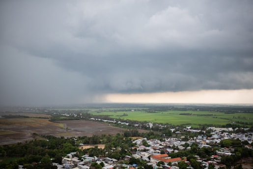 Dark storm clouds loom over a rural area with fields and houses. The clouds are dense and threatening, suggesting an imminent downpour. The landscape below is a mix of green fields and a small settlement with various buildings.