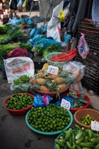 A vibrant market stall displays a variety of fresh produce. There are baskets filled with limes, pineapples, cucumbers, and other leafy greens. Plastic bags, crates, and sacks are scattered around, indicating an active trading area. Price tags are visible on some products.