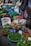 A vibrant market stall displays a variety of fresh produce. There are baskets filled with limes, pineapples, cucumbers, and other leafy greens. Plastic bags, crates, and sacks are scattered around, indicating an active trading area. Price tags are visible on some products.