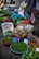 A vibrant market stall displays a variety of fresh produce. There are baskets filled with limes, pineapples, cucumbers, and other leafy greens. Plastic bags, crates, and sacks are scattered around, indicating an active trading area. Price tags are visible on some products.
