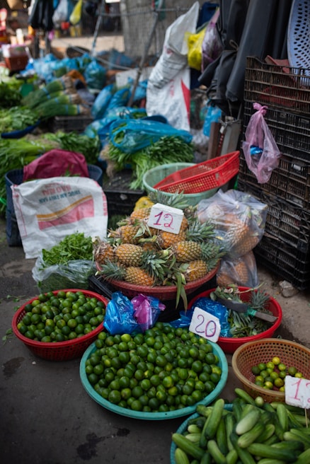 A vibrant market stall displays a variety of fresh produce. There are baskets filled with limes, pineapples, cucumbers, and other leafy greens. Plastic bags, crates, and sacks are scattered around, indicating an active trading area. Price tags are visible on some products.