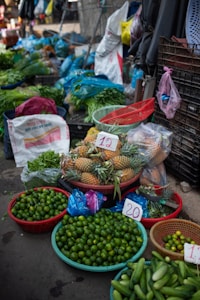 A vibrant market stall displays a variety of fresh produce. There are baskets filled with limes, pineapples, cucumbers, and other leafy greens. Plastic bags, crates, and sacks are scattered around, indicating an active trading area. Price tags are visible on some products.