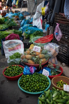 A vibrant market stall displays a variety of fresh produce. There are baskets filled with limes, pineapples, cucumbers, and other leafy greens. Plastic bags, crates, and sacks are scattered around, indicating an active trading area. Price tags are visible on some products.