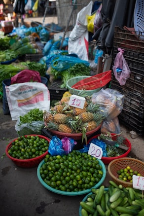A vibrant market stall displays a variety of fresh produce. There are baskets filled with limes, pineapples, cucumbers, and other leafy greens. Plastic bags, crates, and sacks are scattered around, indicating an active trading area. Price tags are visible on some products.