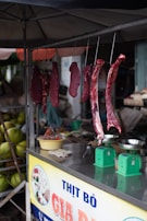 Several cuts of raw meat are hanging from hooks in a small market stall. Below the hanging meat, there are green weighing scales and a yellow bowl on a metallic counter. The stall is covered, providing shade to the items beneath. In the background, there are green coconuts stacked together.