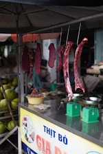 Several cuts of raw meat are hanging from hooks in a small market stall. Below the hanging meat, there are green weighing scales and a yellow bowl on a metallic counter. The stall is covered, providing shade to the items beneath. In the background, there are green coconuts stacked together.