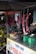 Several cuts of raw meat are hanging from hooks in a small market stall. Below the hanging meat, there are green weighing scales and a yellow bowl on a metallic counter. The stall is covered, providing shade to the items beneath. In the background, there are green coconuts stacked together.