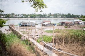 A rural riverside scene with several small houses and huts near the water. A long wooden walkway stretches across grassy and muddy areas leading to some of the structures. The river is calm, with more buildings visible across the water under a cloudy sky. Trees frame the image on the left and right.
