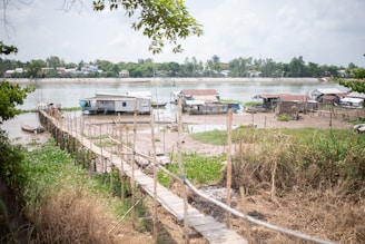 A rural riverside scene with several small houses and huts near the water. A long wooden walkway stretches across grassy and muddy areas leading to some of the structures. The river is calm, with more buildings visible across the water under a cloudy sky. Trees frame the image on the left and right.