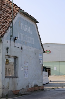 The image features an old building with a worn-down exterior. The walls are painted in faded blue, with visible signs of peeling and weathering. The building's roof is made of weathered red tiles. On the side of the building, there's a large faded sign with the text 'Farma Uherčice'. Various small signs are posted near a small window. In the background, a modern industrial building is visible with stacks of materials and a loading area.