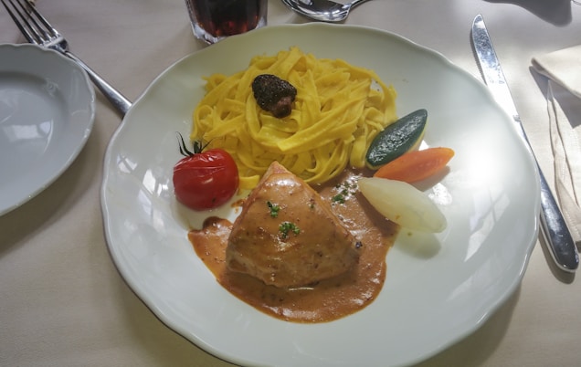 A white plate contains a serving of creamy pasta garnished with a small mushroom on top, accompanied by a piece of chicken bathed in a rich brown sauce. On the side, there is a bright red cherry tomato, and cooked vegetables including a slice of zucchini, a carrot, and an onion. Cutlery is placed on either side of the plate, and a glass of dark beverage is partially visible in the background.