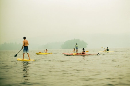 Several people are engaging in water activities on a calm lake. A person on a paddleboard is standing and paddling, while others are kayaking or sitting in small boats. The backdrop features a misty or foggy atmosphere with an indistinct view of distant trees or land.