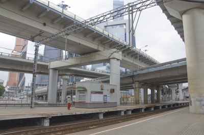 An urban transit setting with multiple overpasses and tall modern skyscrapers in the background. The scene includes a railway platform with tracks, a small shelter for waiting passengers, and a few people scattered around. The architecture is characterized by concrete and metal, typical of urban infrastructure.