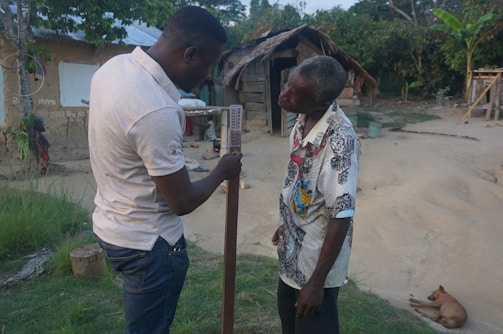 Two men are interacting outside a rustic, thatched-roof structure, possibly in a rural setting. One man is holding a long stick or measuring instrument. The area around them is sandy with some patches of grass, and there is a small dog lying on the ground nearby. Trees and greenery surround the backdrop.