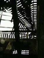 A dilapidated wooden roof structure with gaps and missing planks allowing light to filter through. The beams appear weathered and are casting shadows on the interior walls of the building. Brick walls are partially visible with sections of peeling plaster adding to the aged appearance.