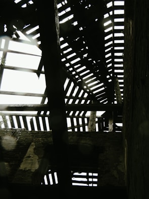 A dilapidated wooden roof structure with gaps and missing planks allowing light to filter through. The beams appear weathered and are casting shadows on the interior walls of the building. Brick walls are partially visible with sections of peeling plaster adding to the aged appearance.